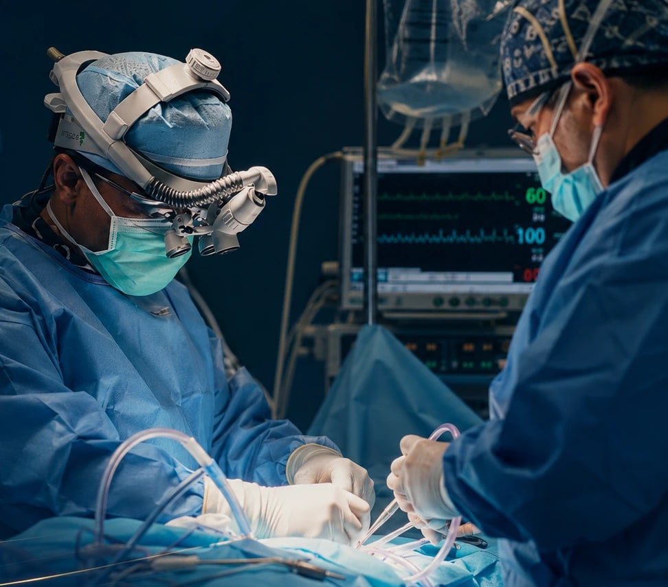 Two surgeons operating under magnification loupes beside a patient monitor